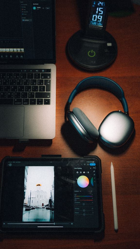 Top view of a laptop, headphones, tablet, and stylus on a wooden desk.